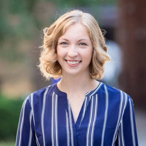 A smiling woman with short blonde curly hair wearing a blue and white striped blouse, standing outdoors with a blurred background.